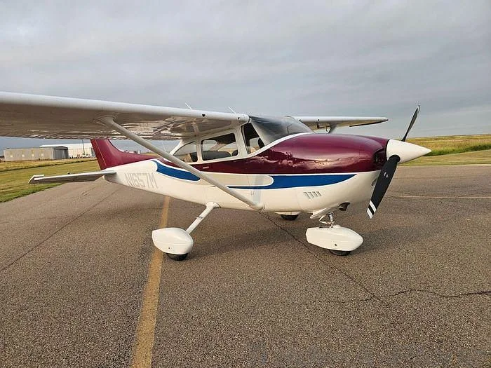A white and maroon Cessna 182 Skylane with blue accents, parked on an airfield under an overcast sky.