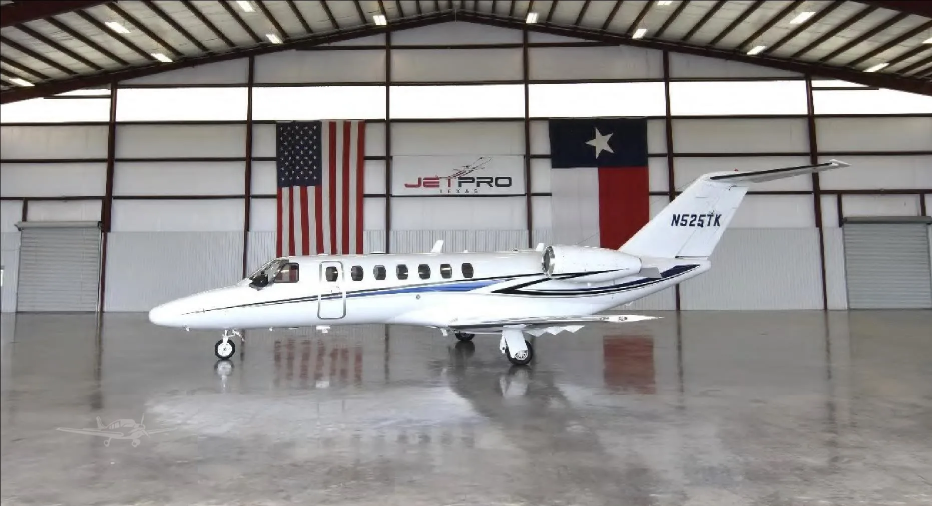A white Cessna CitationJet with blue and black stripes parked in a hangar with US and Texas flags.