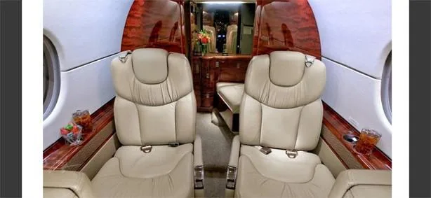 A luxurious aircraft cabin with two beige leather seats facing forward, dark wood paneling, and drinks on side tables.