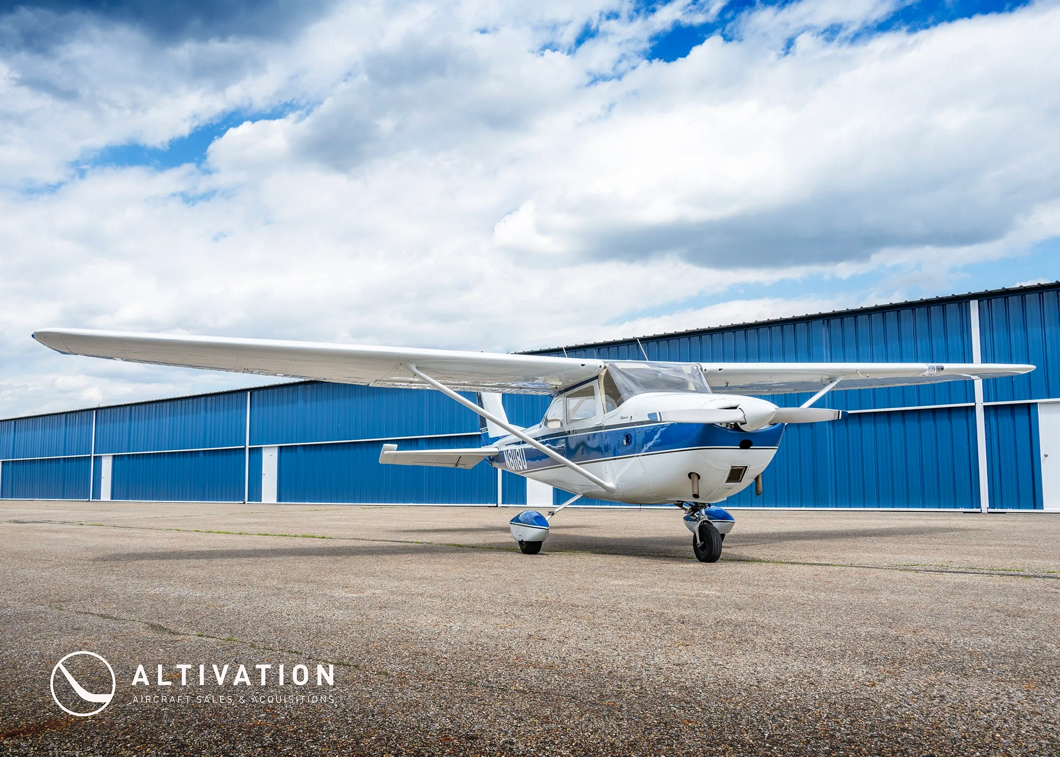 A low-angle front view of the white and blue Cessna 172 Skyhawk N8118U parked on an asphalt ramp in front of a long blue hangar.