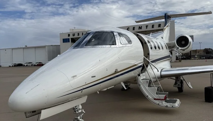 A close-up front view of a white Embraer Phenom 300 private jet with its passenger door open and stairs extended, parked in front of a business center.