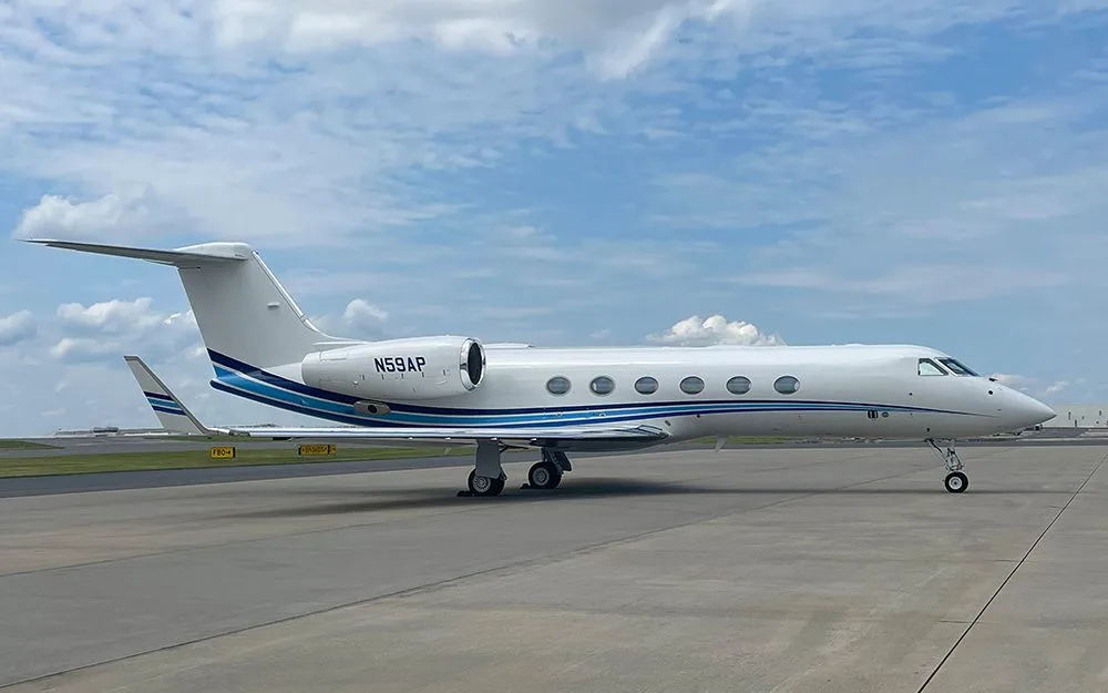 A white Gulfstream G550 business jet with blue and black stripes on the fuselage and tail, parked on an airport tarmac under a partly cloudy sky.