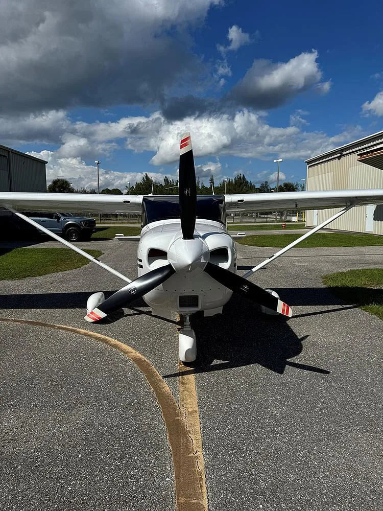 Head-on view of a white Cessna 182 Skylane with black propeller blades, parked on an asphalt ramp.