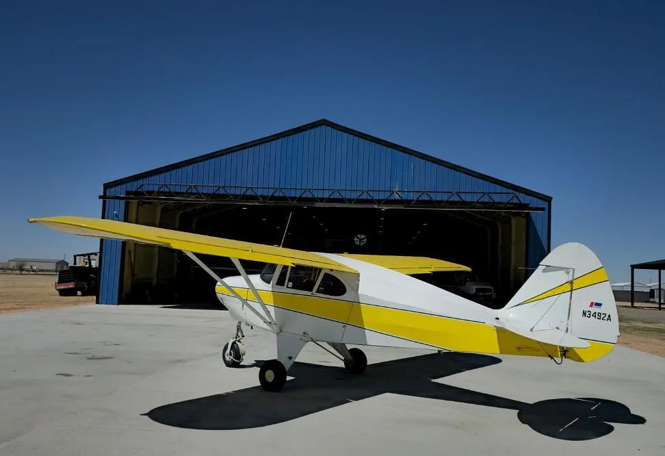 A white and yellow Piper PA-12 Super Cruiser (N3492A) parked in front of a blue hangar.