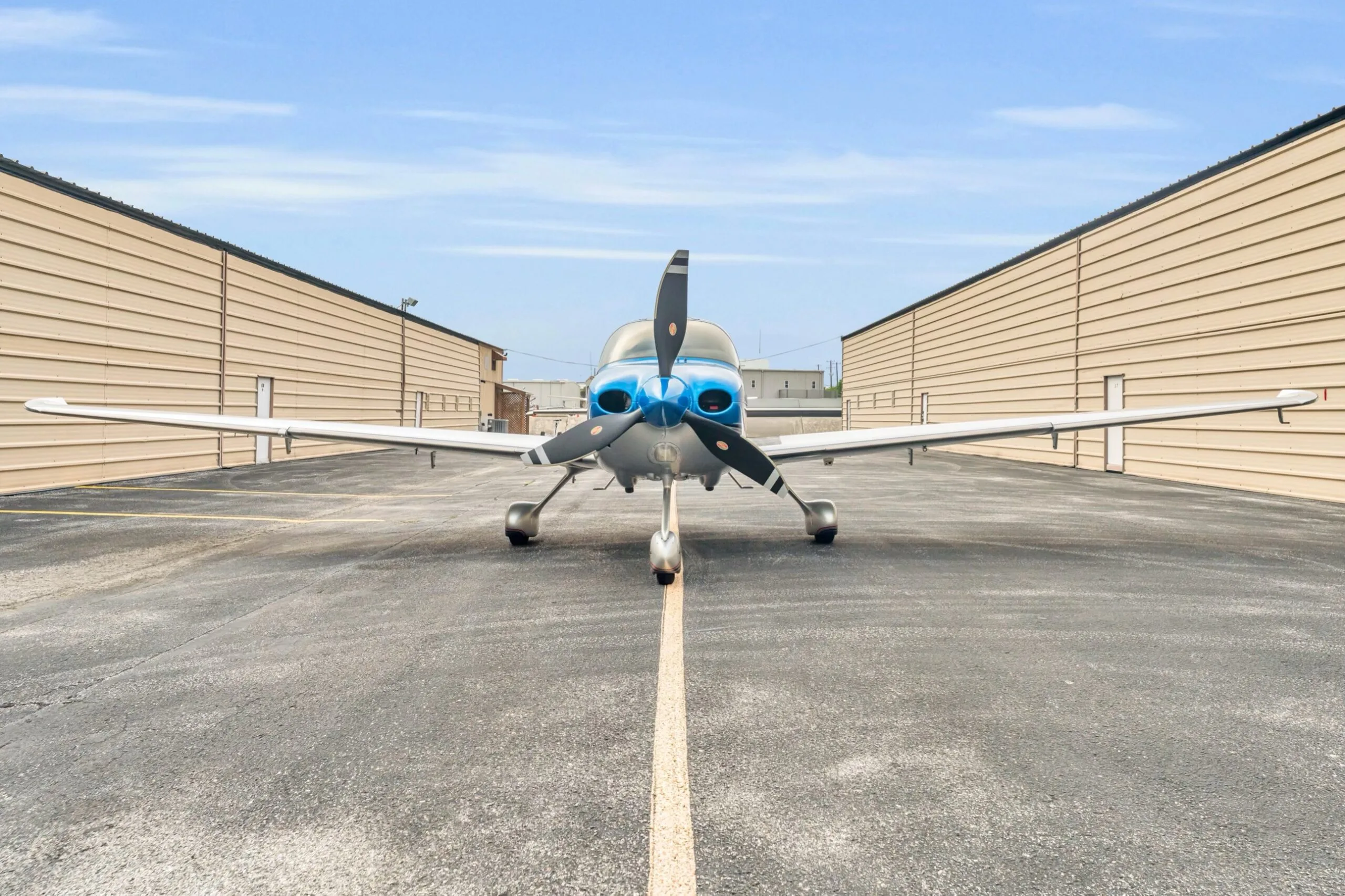 A head-on view of the blue and silver Cirrus SR22T GTS aircraft N66TM, centered on an asphalt taxiway between hangars.
