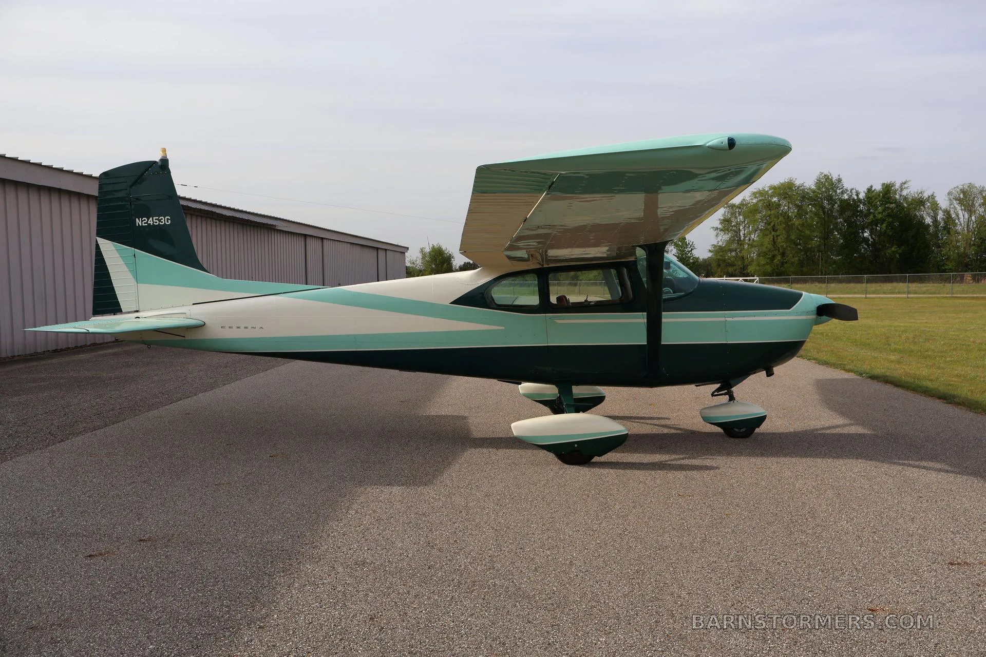A green and white single-engine propeller aircraft (Cessna 172) is parked on an asphalt ramp next to a hangar, viewed from the side.