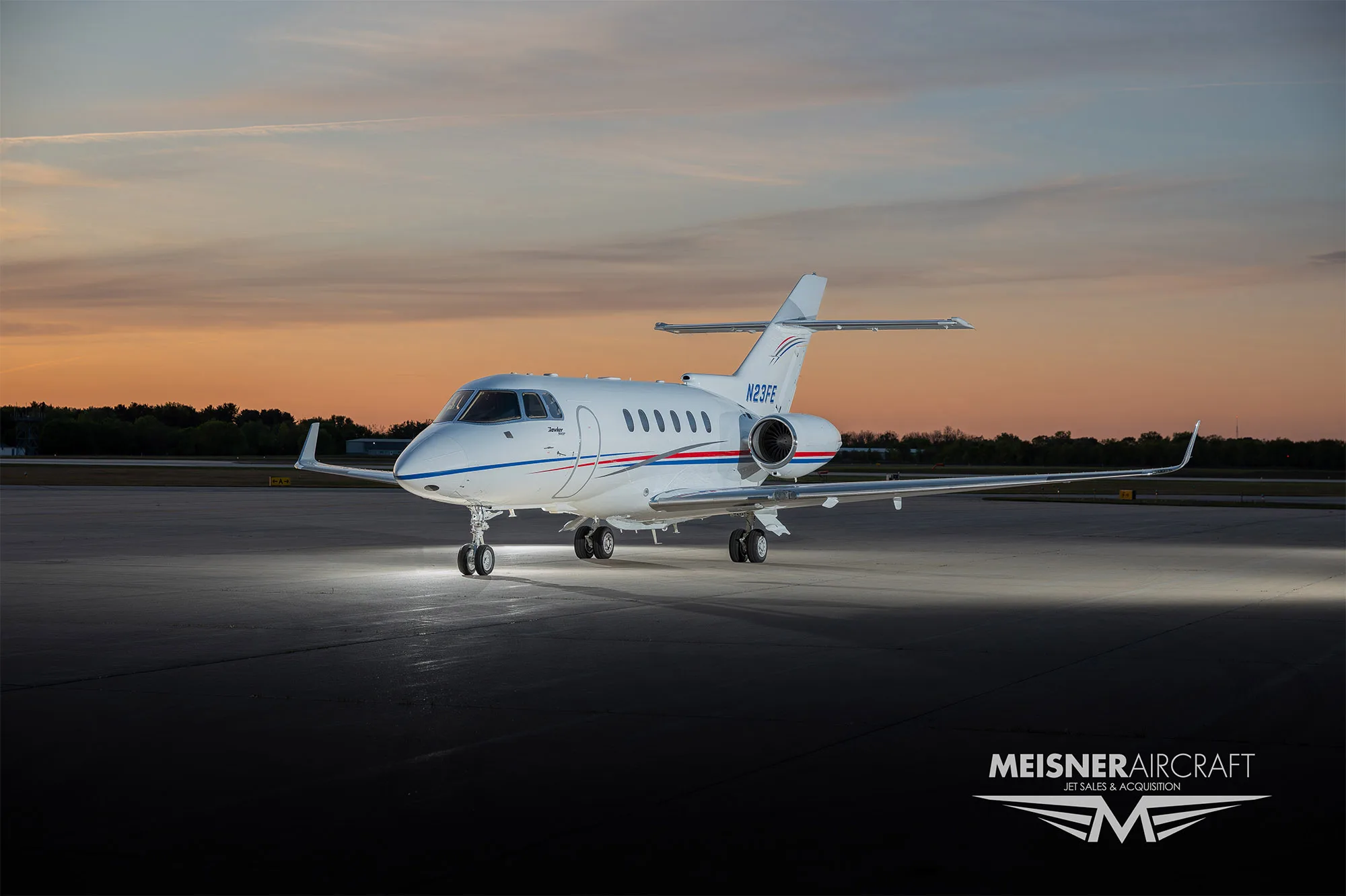 A white business jet with red and blue stripes, registration N23FE, parked on an airfield at sunset, viewed from the front-left.