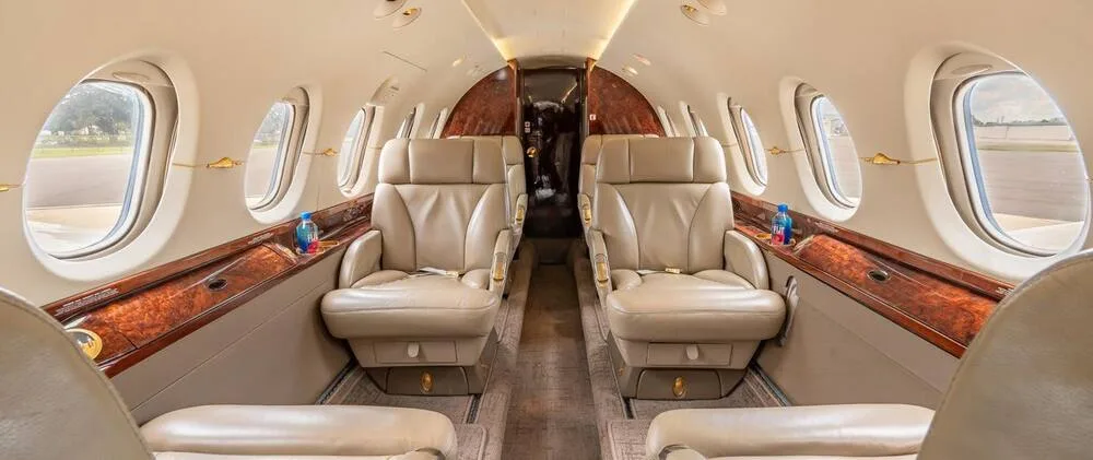 The luxurious cabin interior of a business jet, featuring two rows of beige leather seats and polished wood paneling.