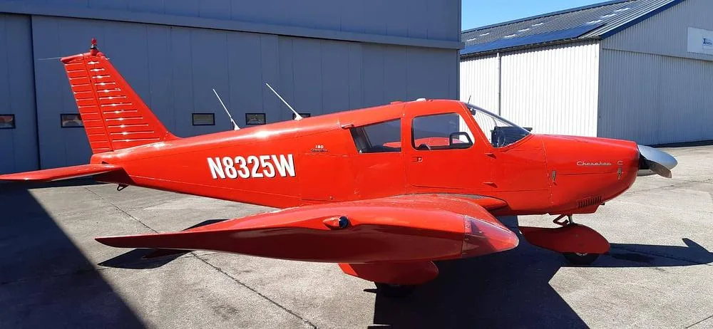 A red Piper Cherokee 180 (N8325W) parked on an tarmac in front of hangars, viewed from the side.