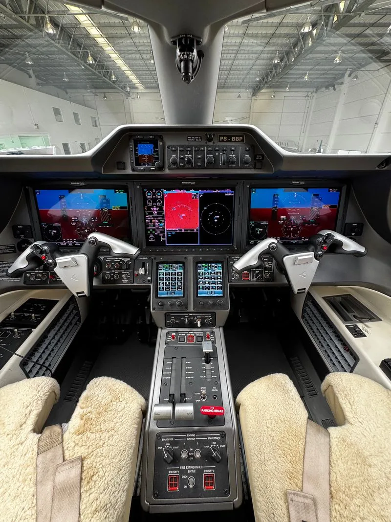 The modern cockpit of an Embraer Phenom 300 with multiple digital displays, controls, and sheepskin seat covers.