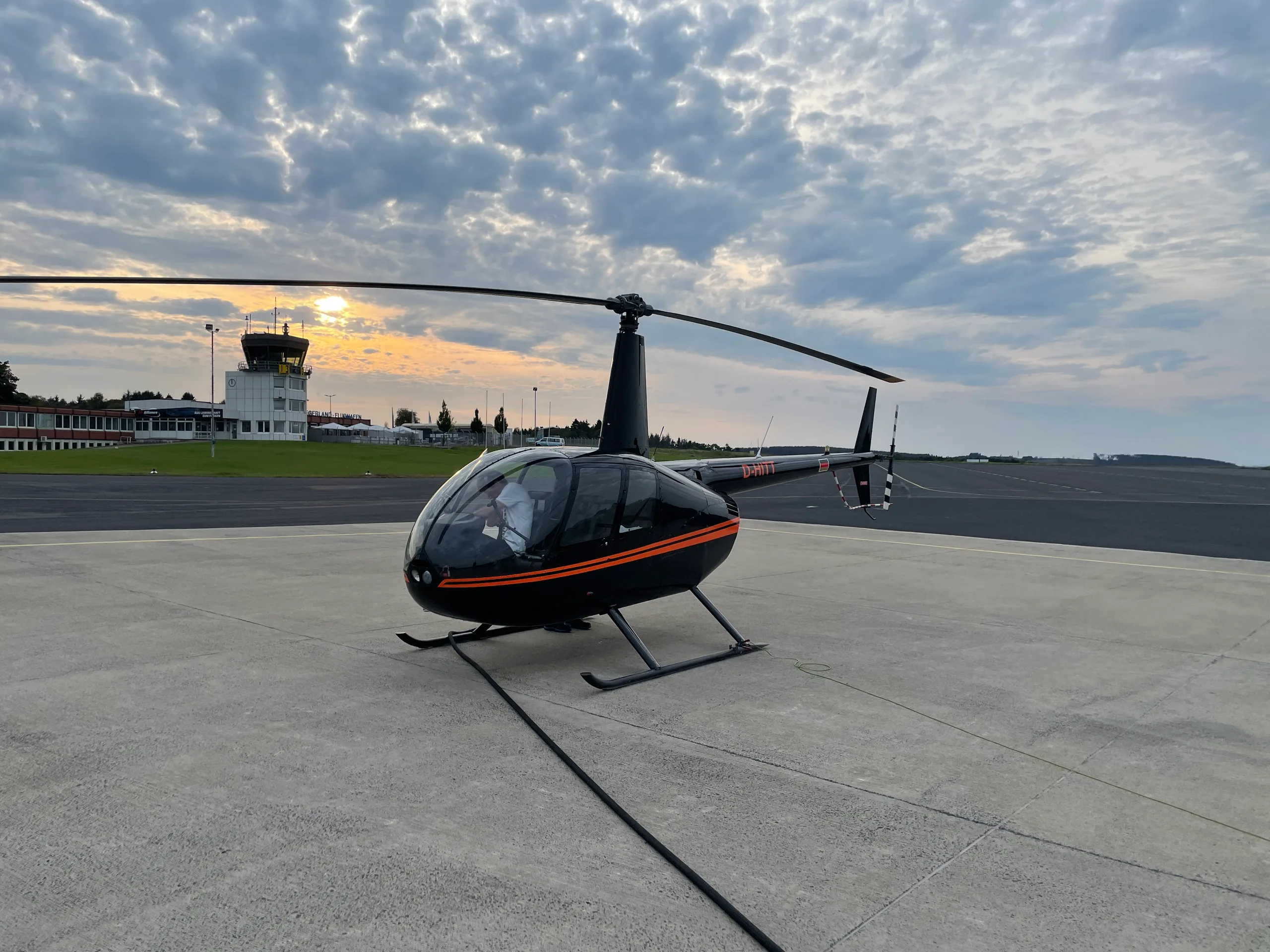 A black Robinson R44 helicopter with orange stripes is parked on an airfield at sunset, with a control tower in the background.