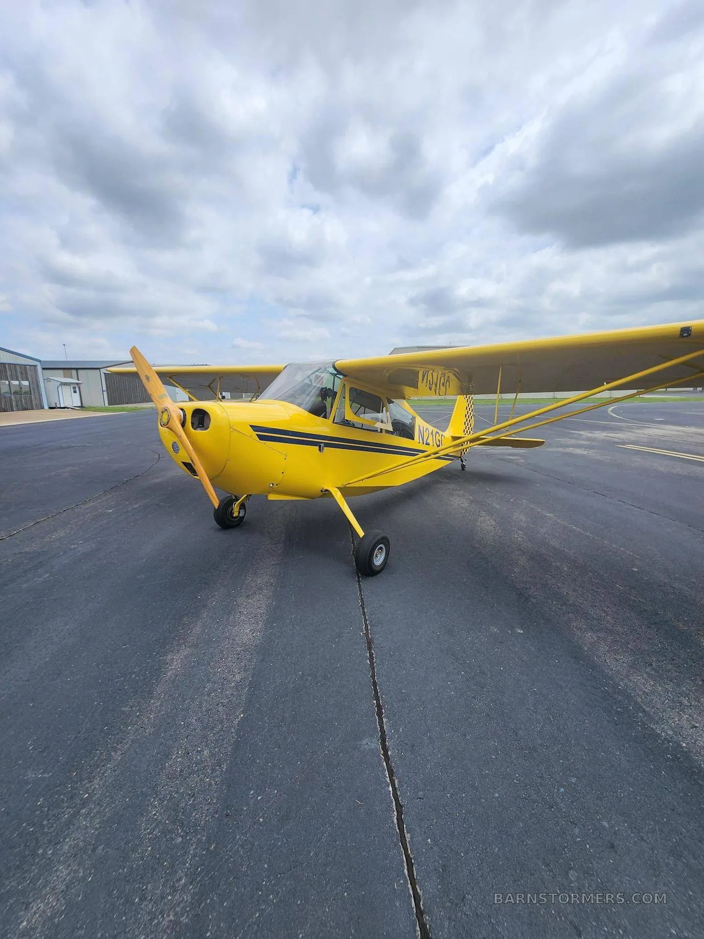A three-quarter front view of a yellow single-engine aircraft with a wooden propeller and black stripe, registration N21GC, parked on an asphalt tarmac.