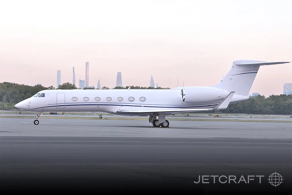 A white Gulfstream G550 business jet with blue stripes on a tarmac with a city skyline in the background at dusk.