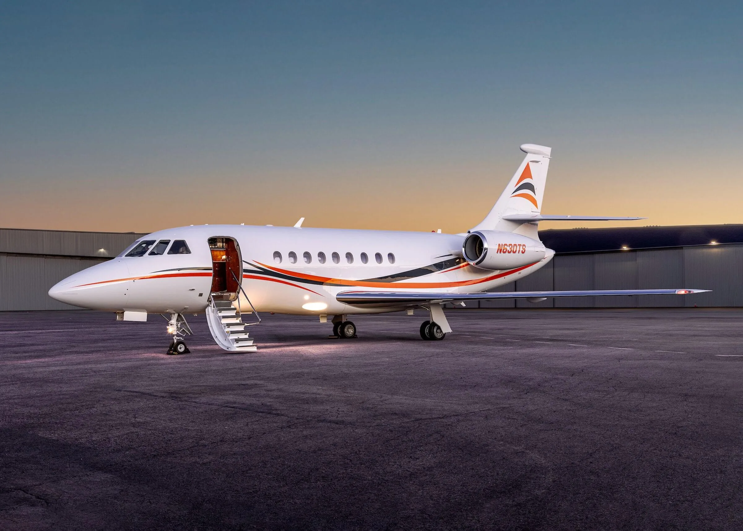 A white Dassault Falcon business jet with orange and black stripes and registration N630TS is parked on an tarmac at dusk, with its main door open.