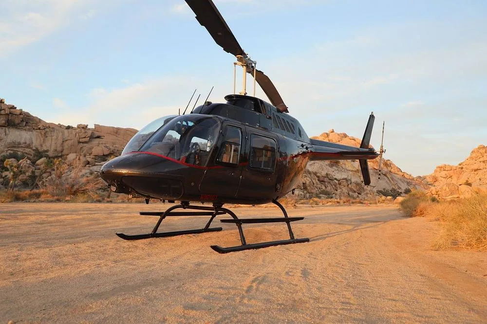 A black helicopter with red trim sits on a dirt landing pad with rocky desert hills in the background.