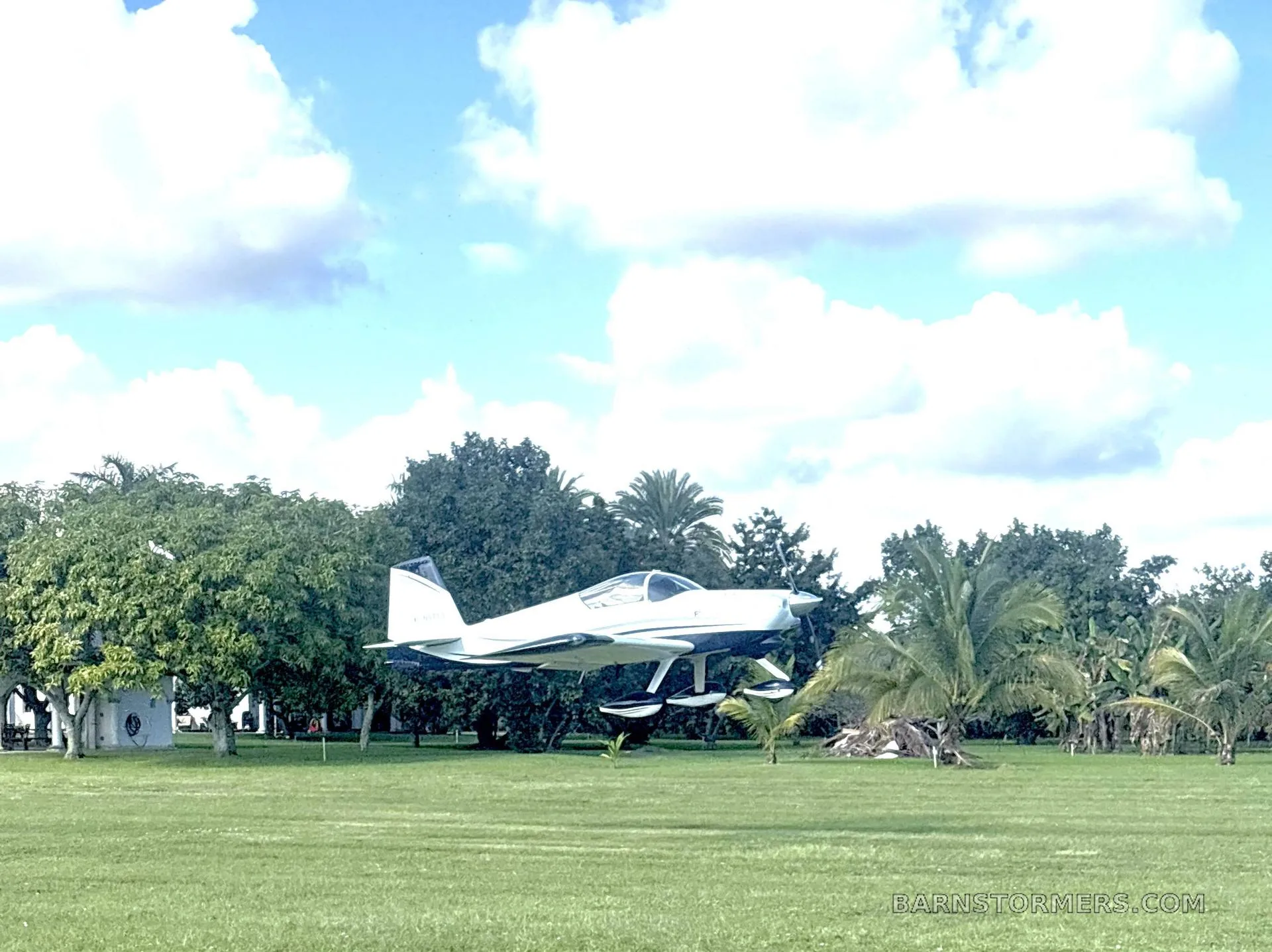 A white and blue Vans RV-7 aircraft with registration N57TG on a grassy field with palm trees under a partly cloudy sky.
