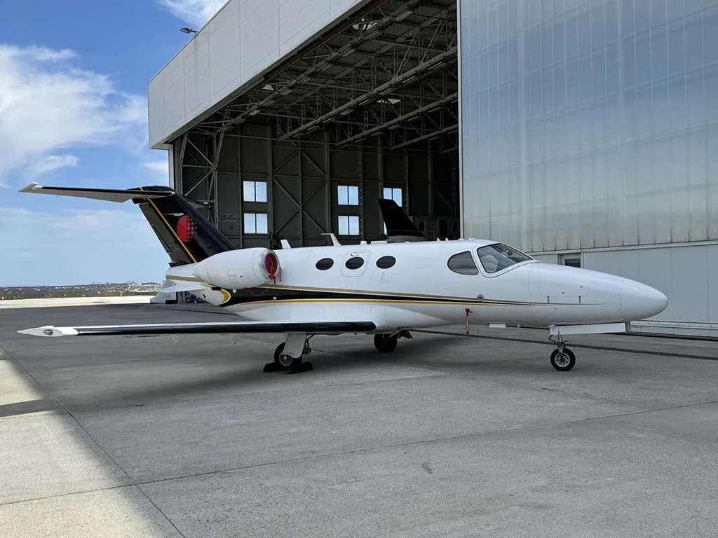 A white business jet with black and gold stripes on the tail, parked on an airport tarmac in front of a modern hangar.