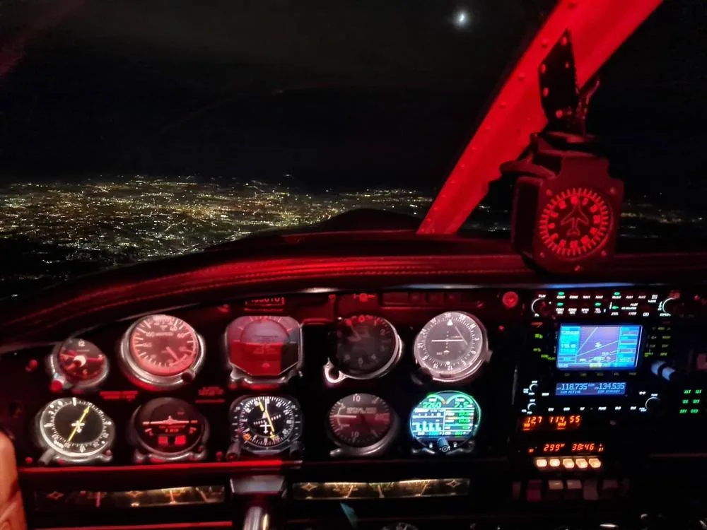 The cockpit of a Piper Archer II at night, showing illuminated instruments and city lights below.
