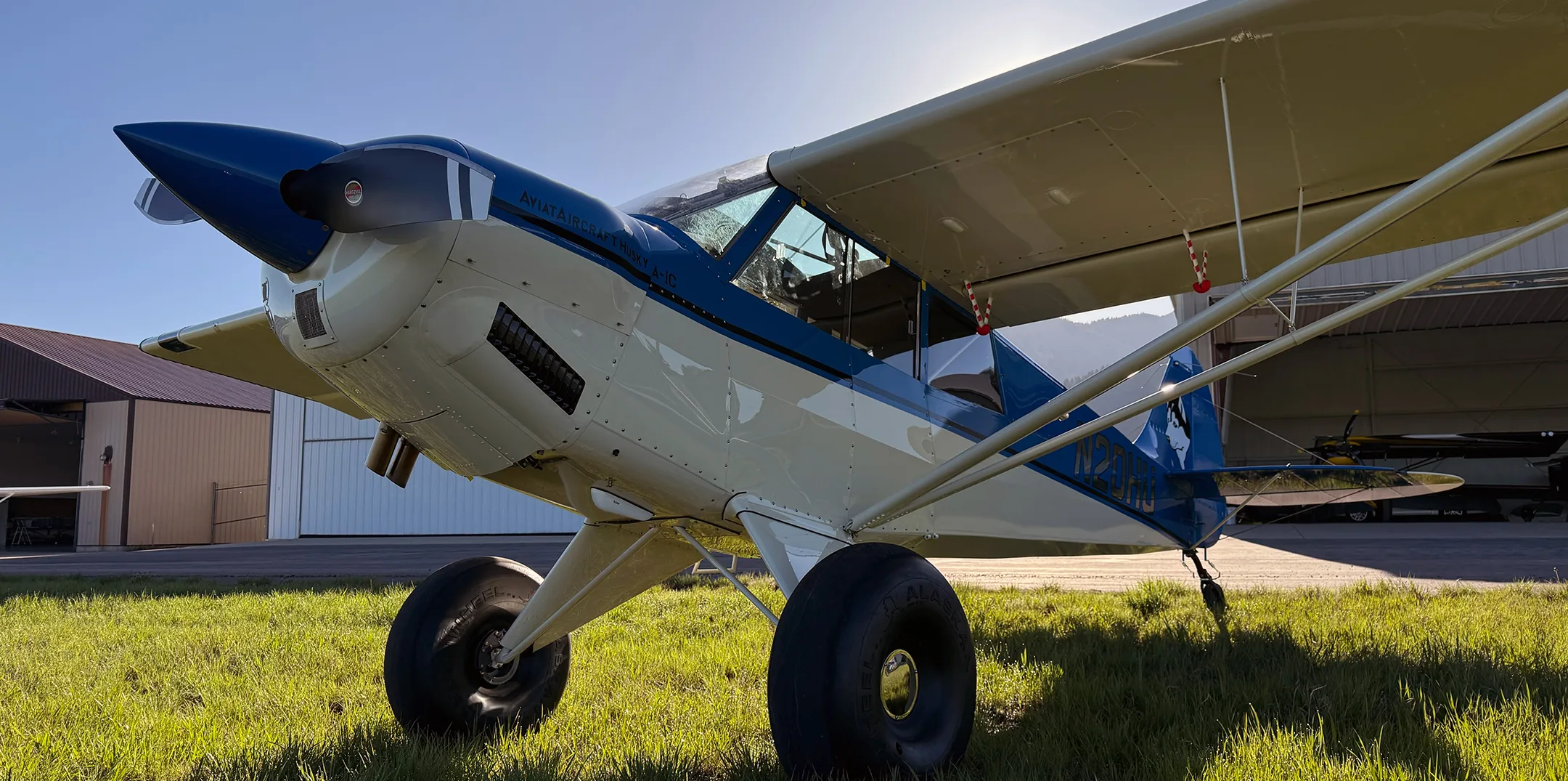 A blue and white Aviata Aircraft Husky A-1C with large tires parked on a grassy field in front of hangars and mountains.