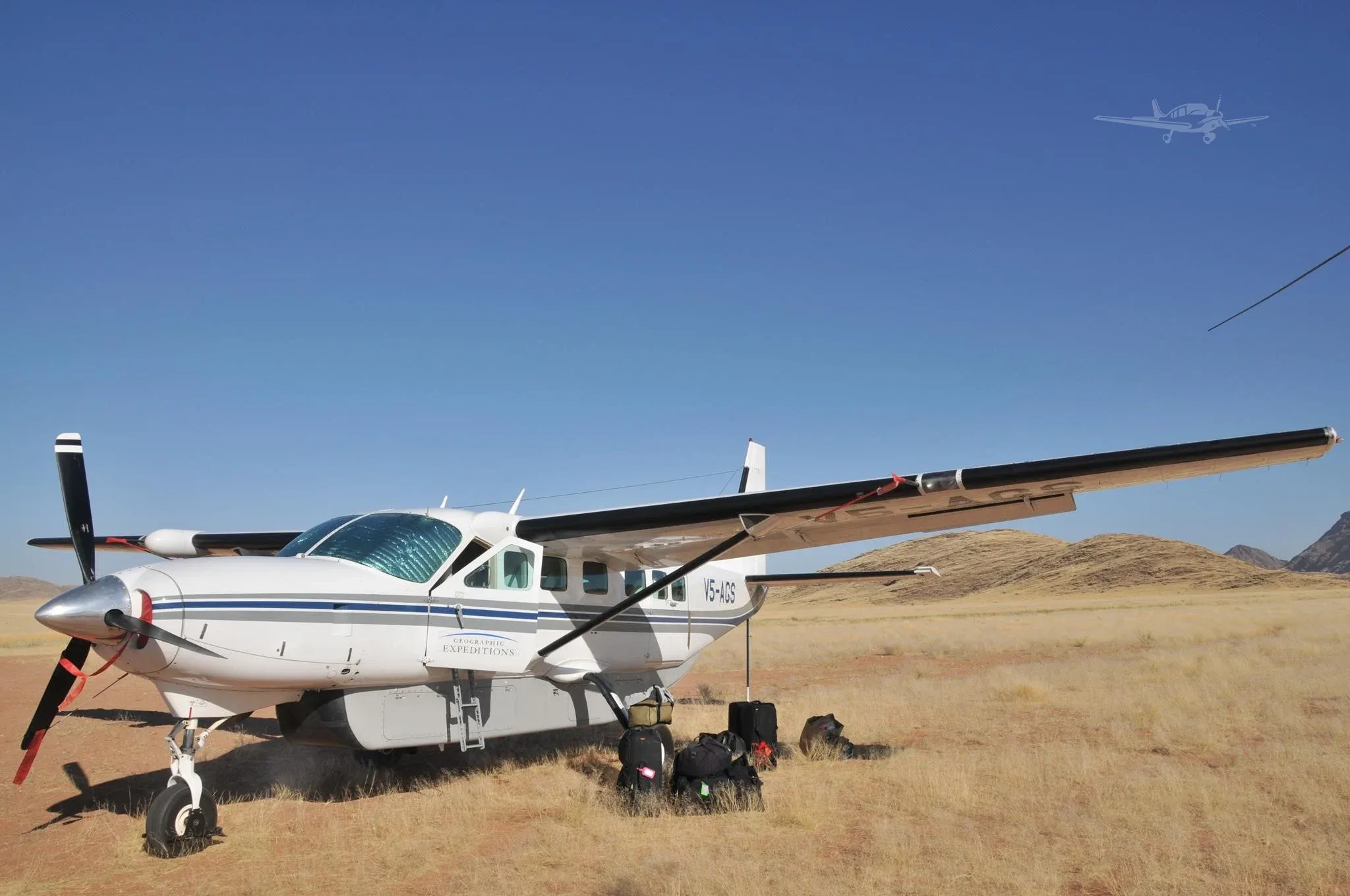 A white Cessna Caravan with blue and grey stripes, registration V5-AGS, is parked on a dry grassy field with luggage next to it, under a clear blue sky.