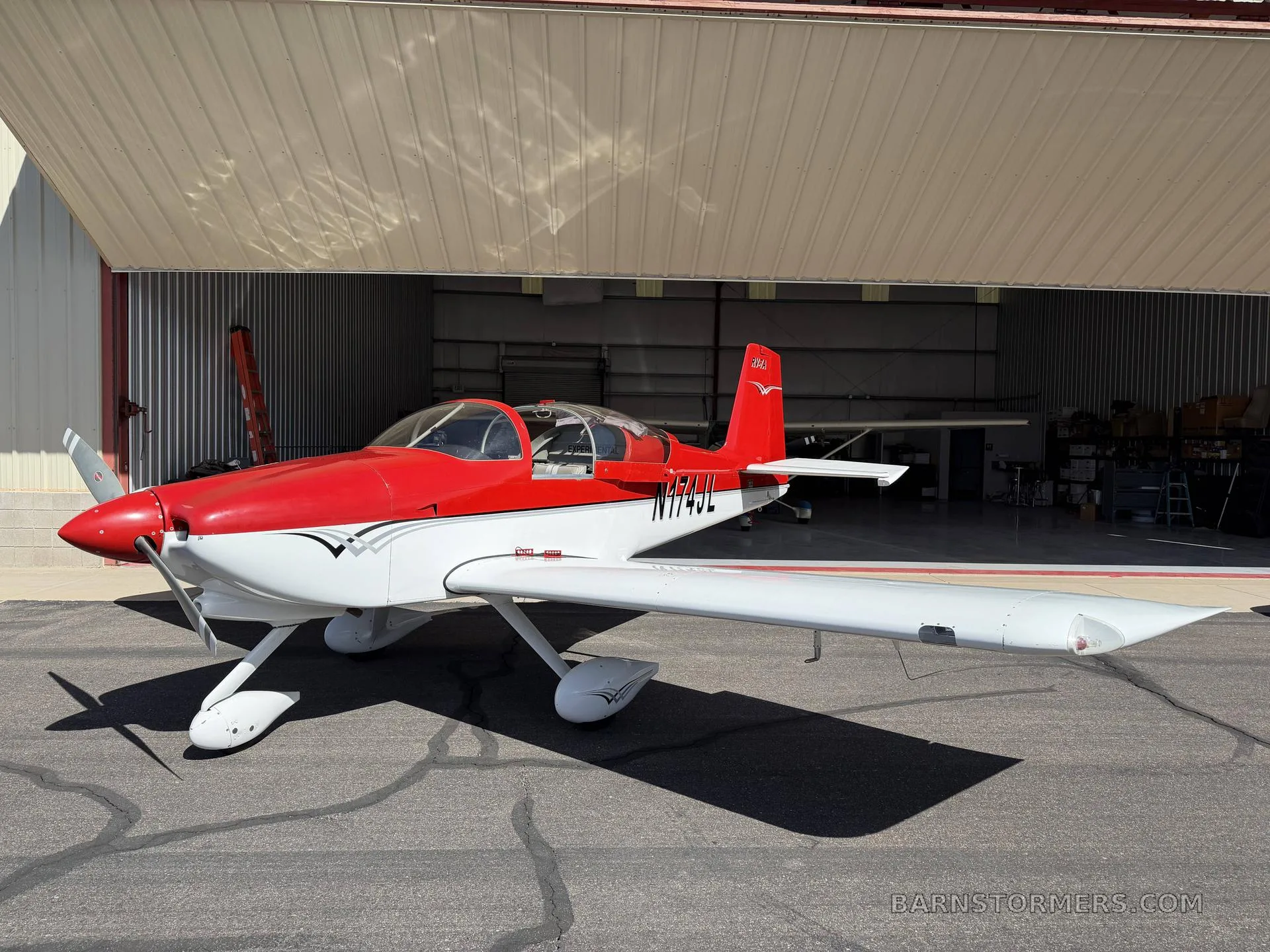 A red and white Vans RV-7A aircraft, registration N174JL, is parked on the tarmac in front of an open hangar, viewed from the side.