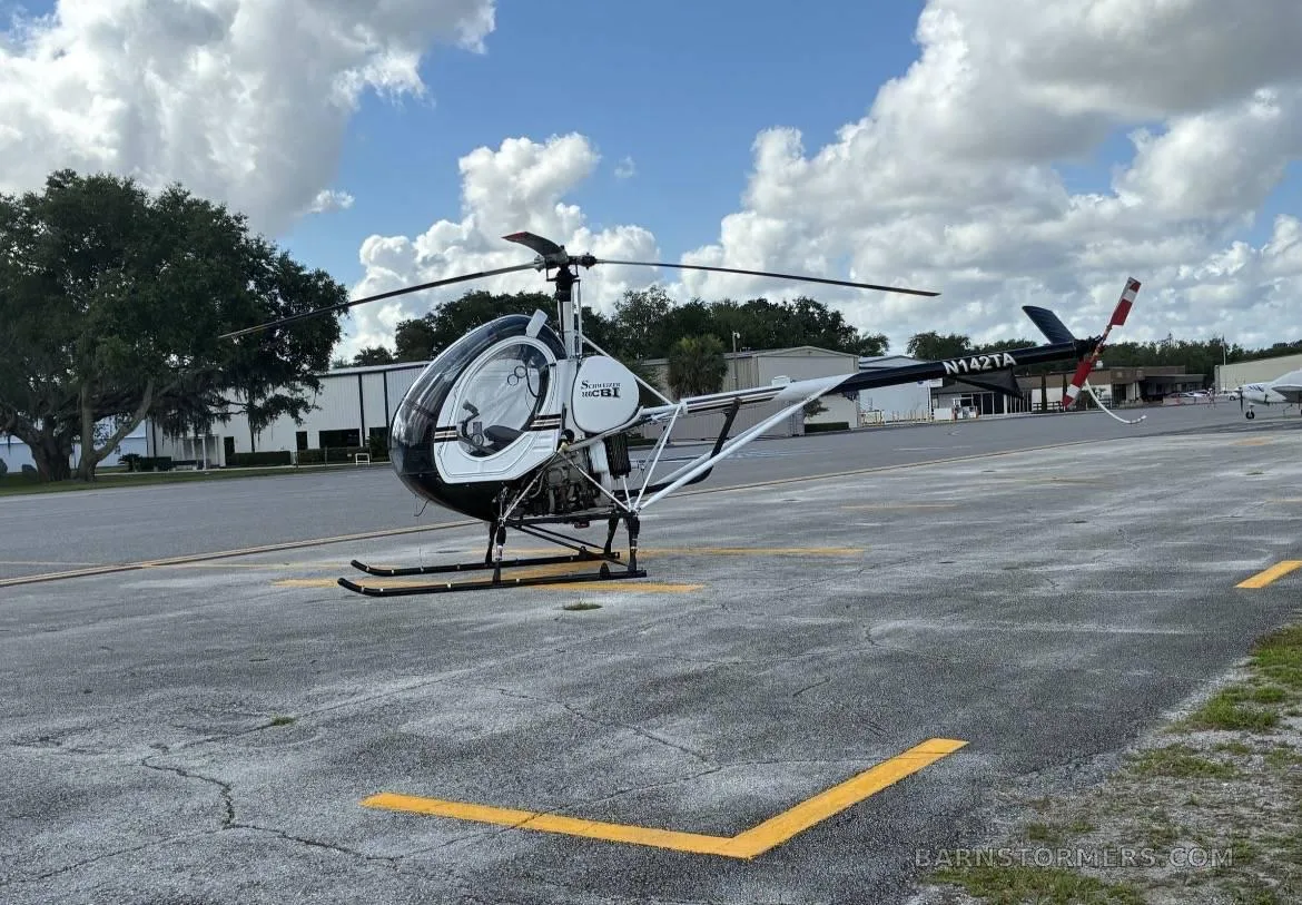 A black and white Schweizer 300CBi helicopter (N142TA) parked on an asphalt tarmac under a partly cloudy sky.