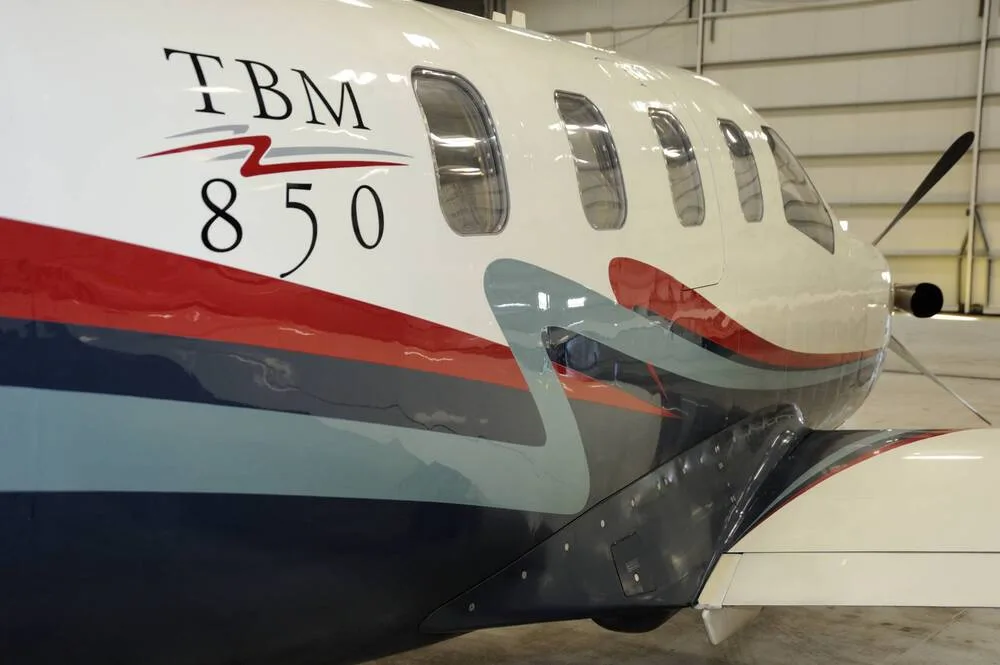 A close-up view of the TBM 850 aircraft's fuselage, showing its white, blue, and red paint scheme and passenger windows.