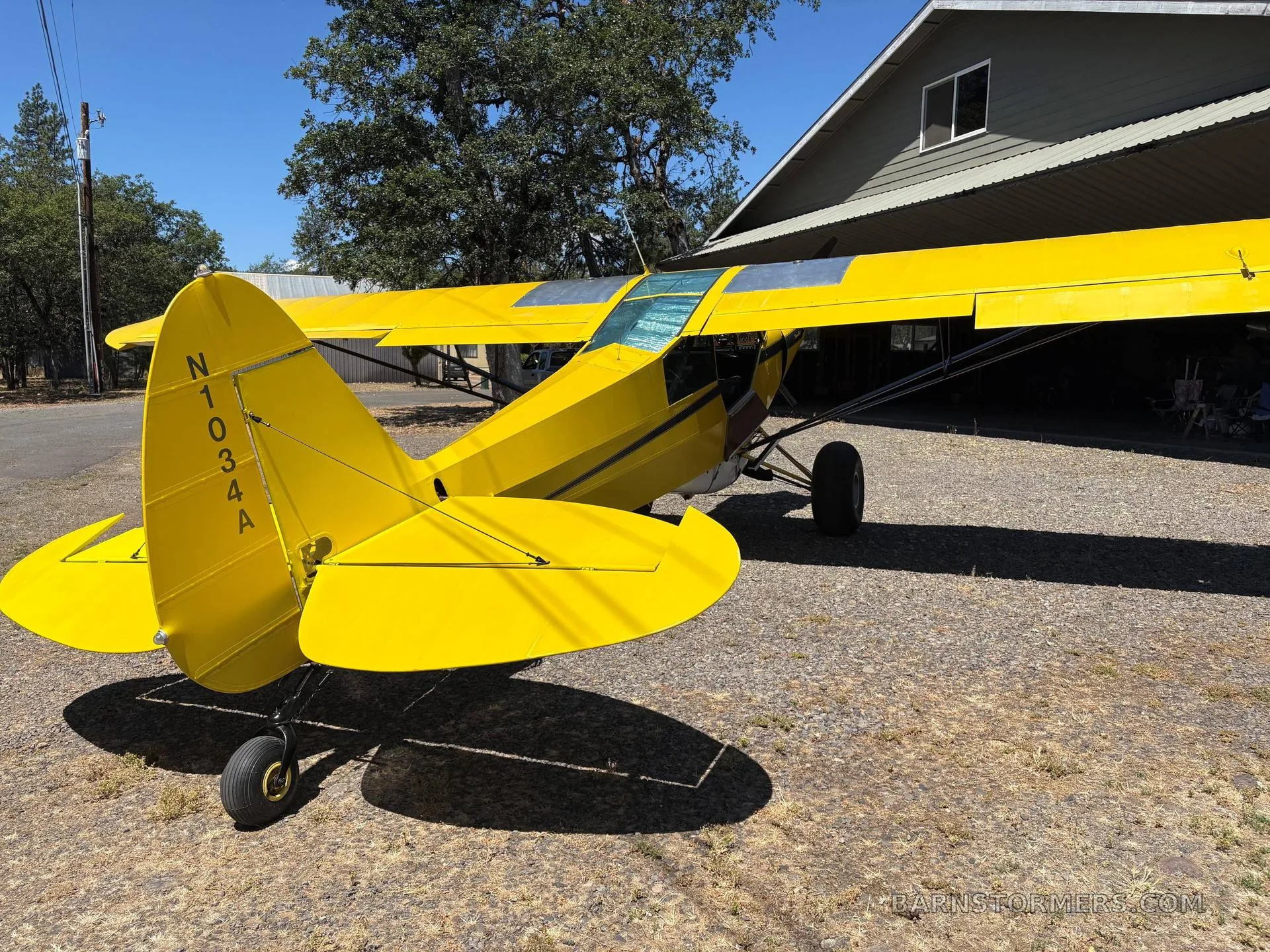 The tail and right wing of a yellow Piper Super Cub with registration N1034A are visible, parked on a gravel surface next to a building.
