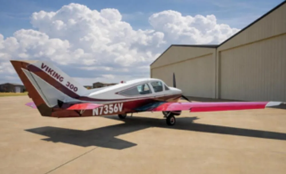A red and white Bellanca Viking 300 (N7356V) parked on an airfield with hangars in the background.