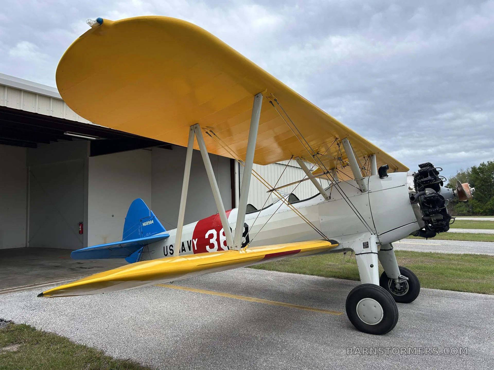 A yellow and white biplane with 'US NAVY 369' markings and a blue tail, parked on an asphalt ramp next to a hangar.