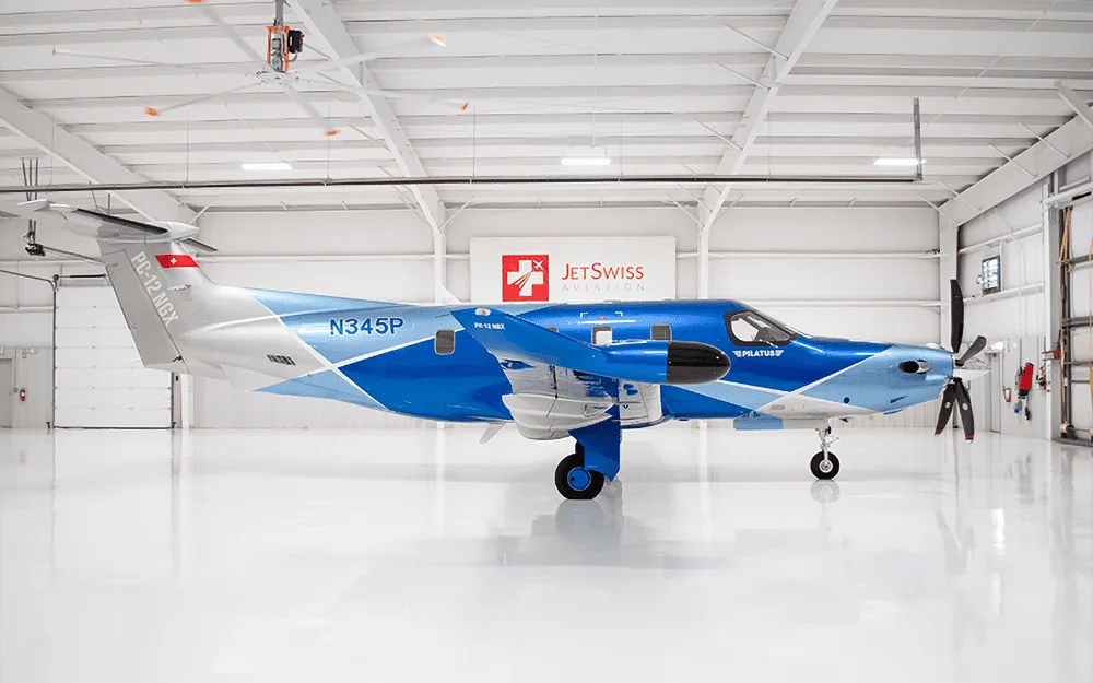A side view of a Pilatus PC-12 NGX aircraft with a blue and silver paint scheme, registration N345P, parked inside a white hangar.