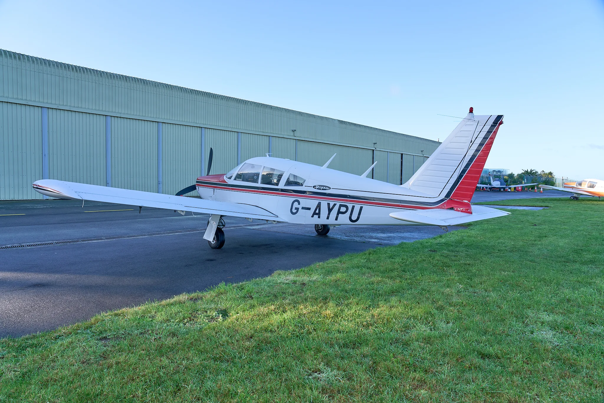 A side view of the white and red Piper Cherokee G-AYPU with a large green hangar in the background.
