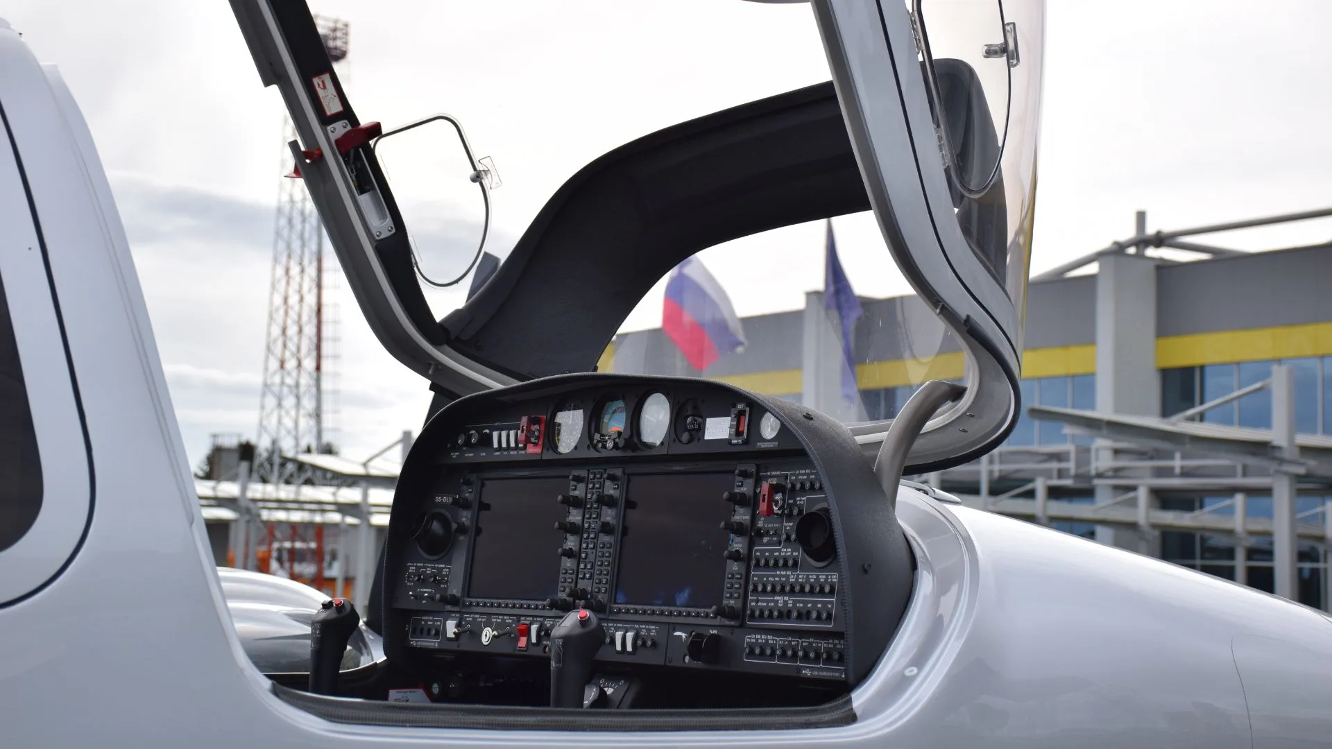 The cockpit of a silver Diamond DA62 aircraft, showing the instrument panel with two large displays and controls, viewed through the open canopy.