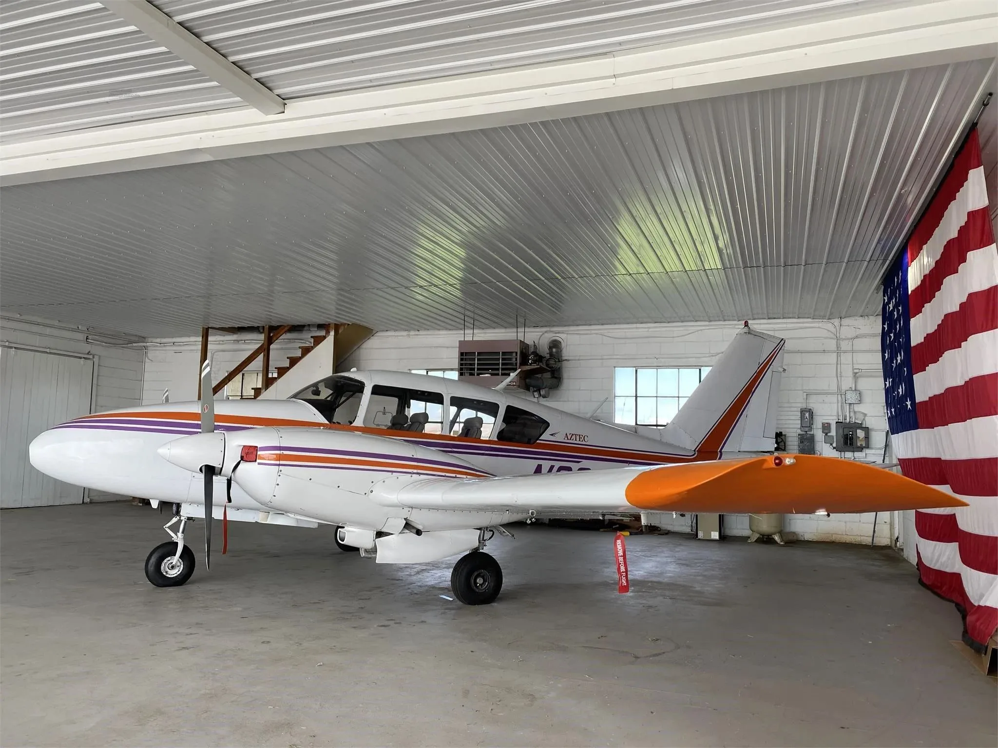 A side view of a white Piper Aztec twin-engine aircraft with orange and purple stripes in a hangar, showing the tail and right wing.