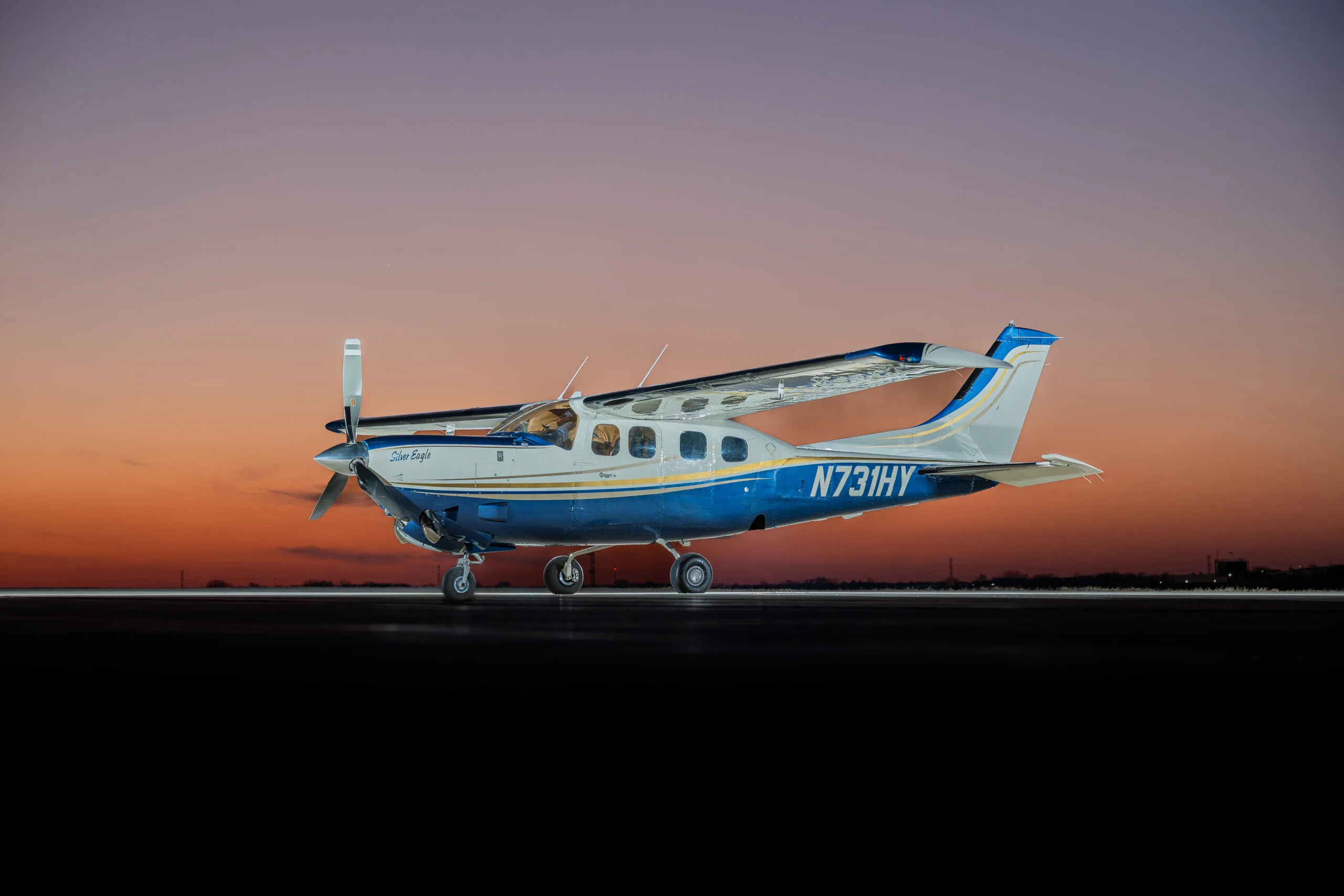 A blue and white Cessna 210 Silver Eagle aircraft with tail number N731HY is parked on a tarmac at sunset.