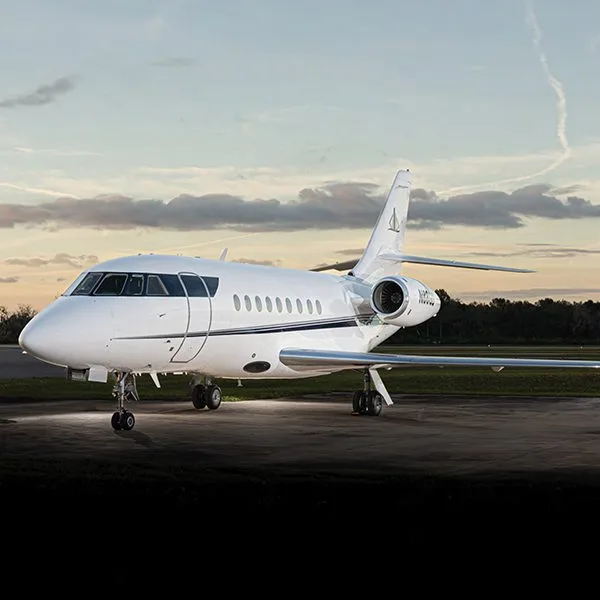 A white Dassault Falcon business jet with a dark stripe on the fuselage sits on a tarmac at sunset.