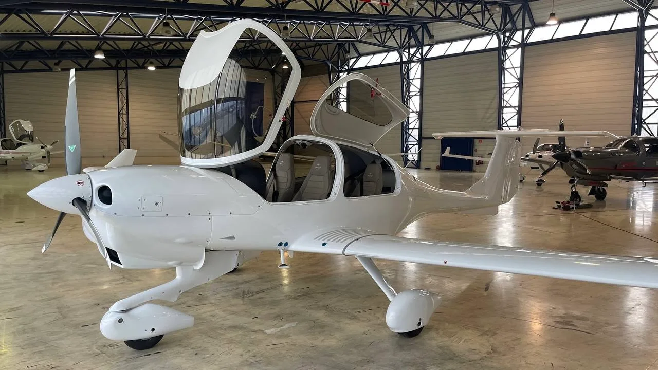 A white Diamond aircraft with its canopy and doors open, parked inside a hangar.