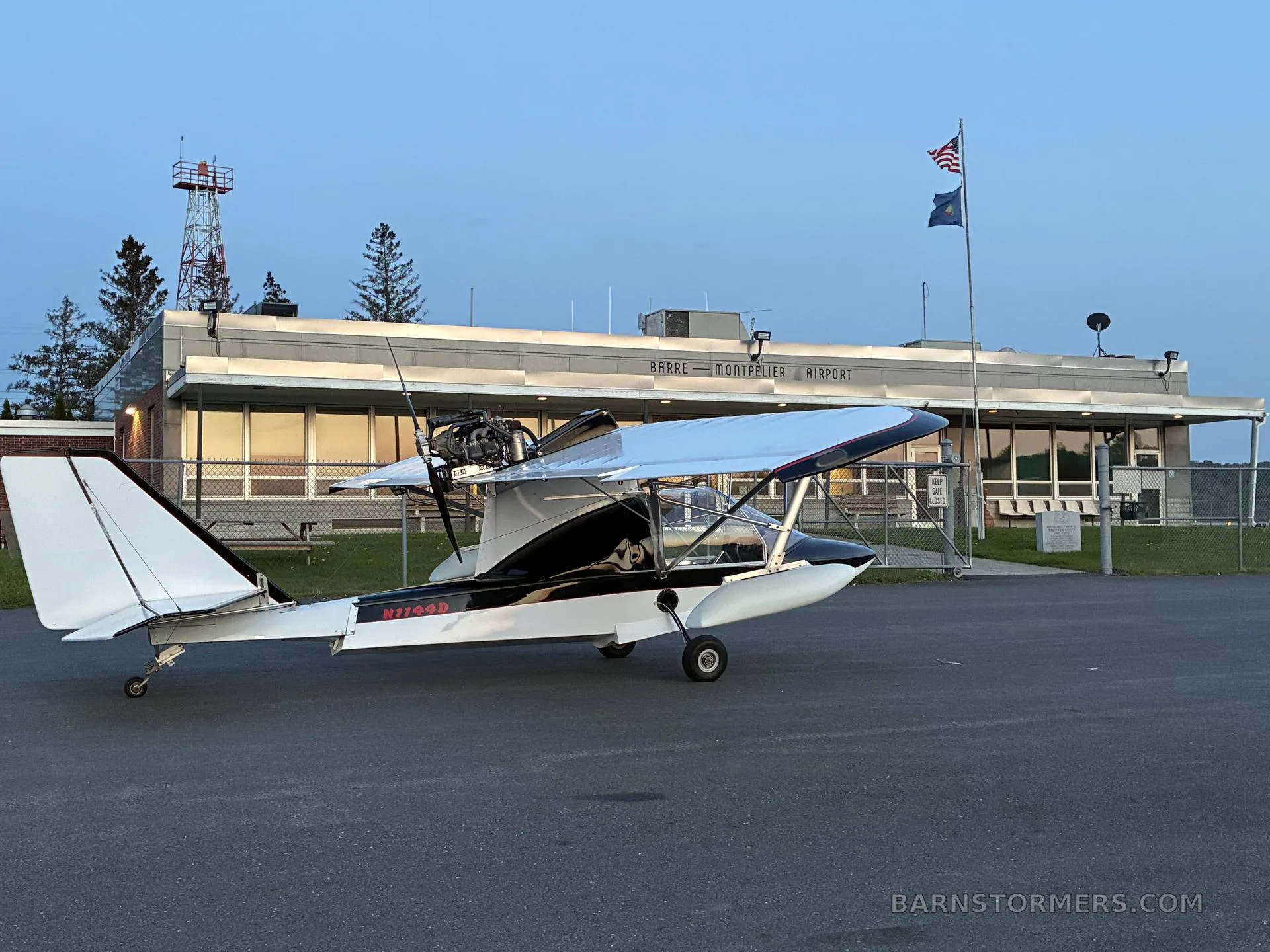 A white and black ultralight aircraft with registration N7749D parked on the tarmac in front of the Barre-Montpelier Airport terminal at dusk.
