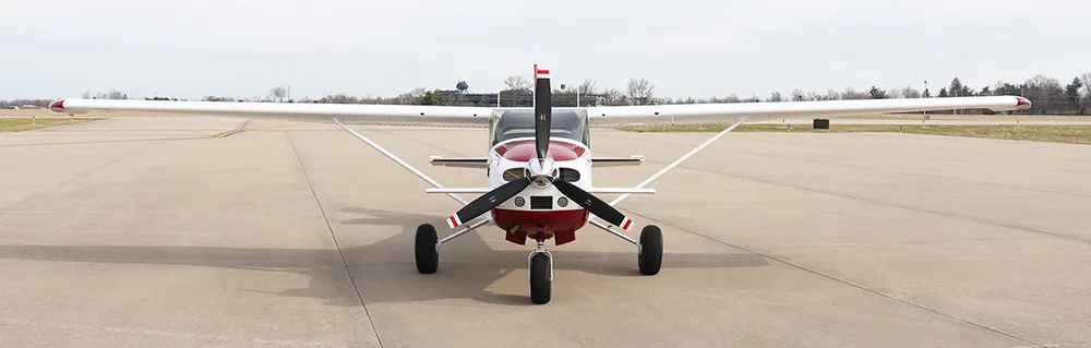 A front view of a white and red single-engine high-wing aircraft with a propeller, parked on an airfield.