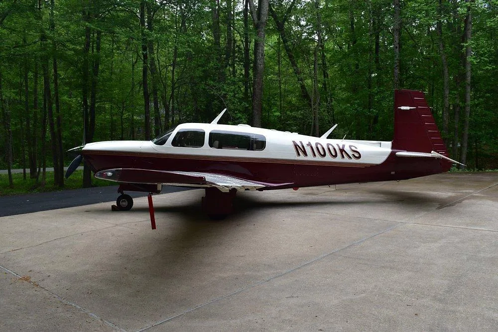 A maroon and white single-engine aircraft, N100KS, parked on a concrete pad with green trees in the background.