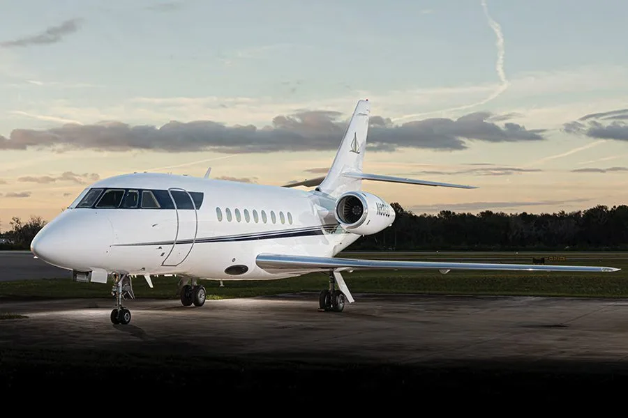 A white Dassault Falcon business jet with a dark stripe on the fuselage is parked on a tarmac at dusk.