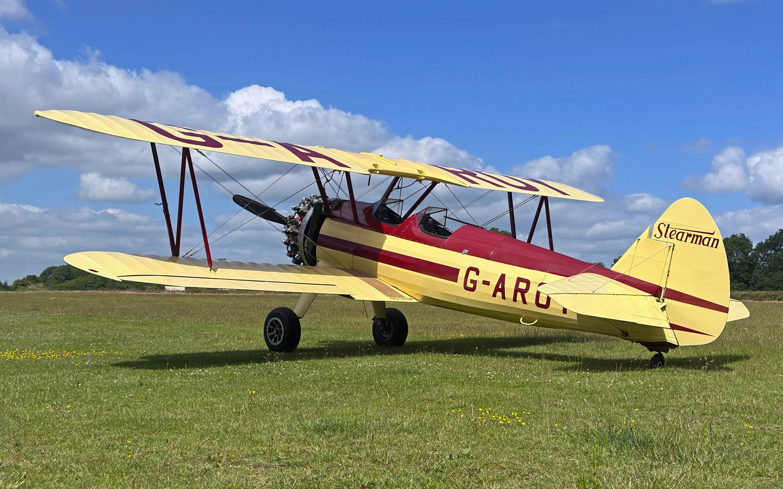 A yellow and red Stearman biplane with registration G-AROY parked on a grassy airfield under a blue sky.