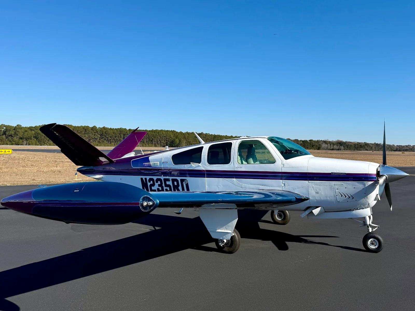 A white Beechcraft Bonanza with purple and blue accents, registration N235RD, parked on an airfield tarmac under a clear blue sky.