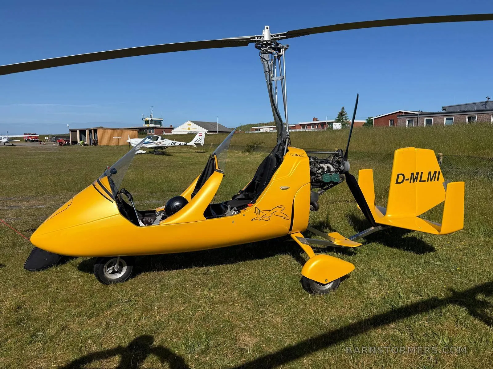 A three-quarter view of a yellow MTO Sport autogyro (D-MLMI) with an open cockpit, parked on a grassy airfield.