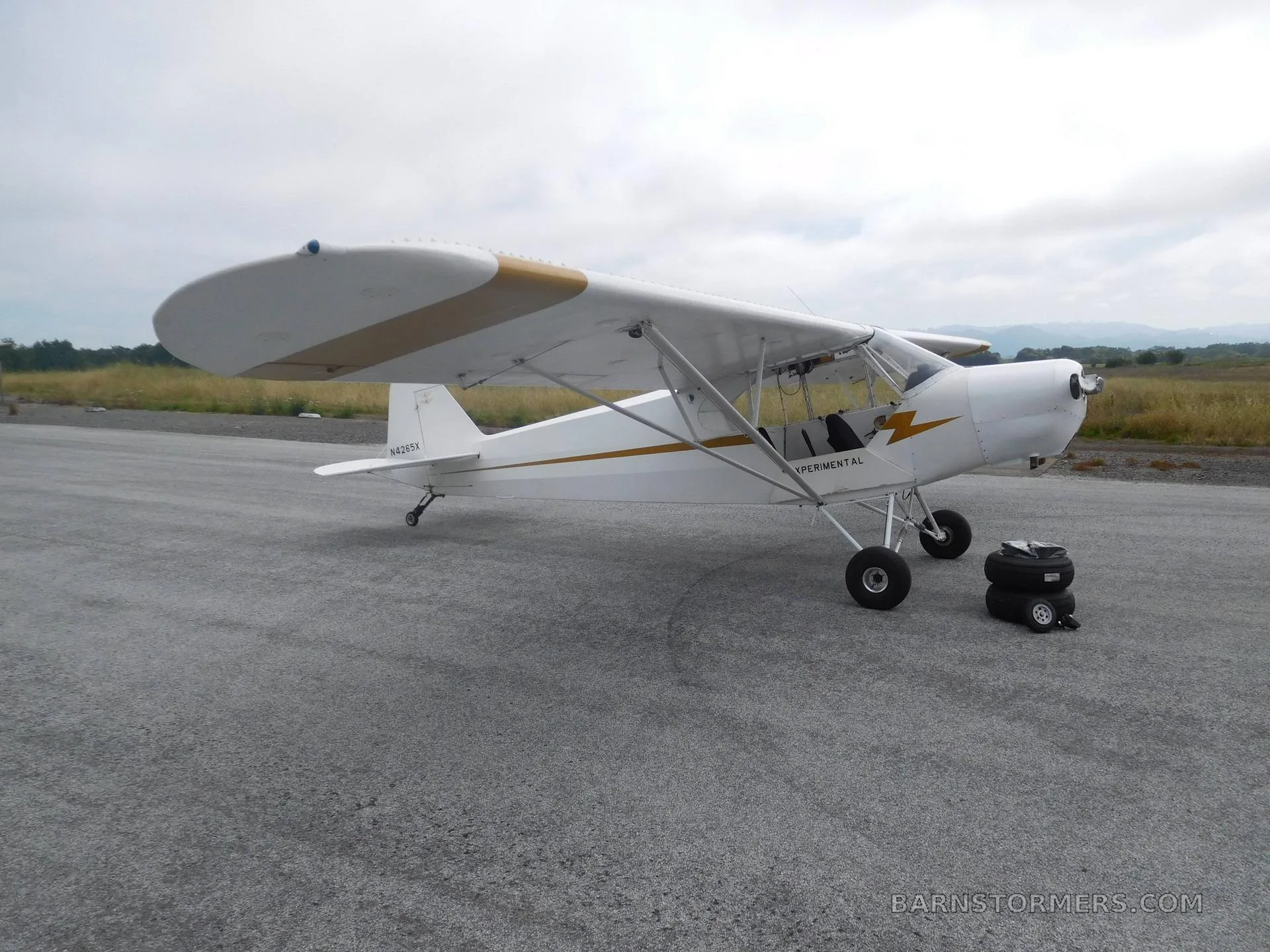 A white experimental aircraft with gold wing stripes is parked on an asphalt runway under a cloudy sky.