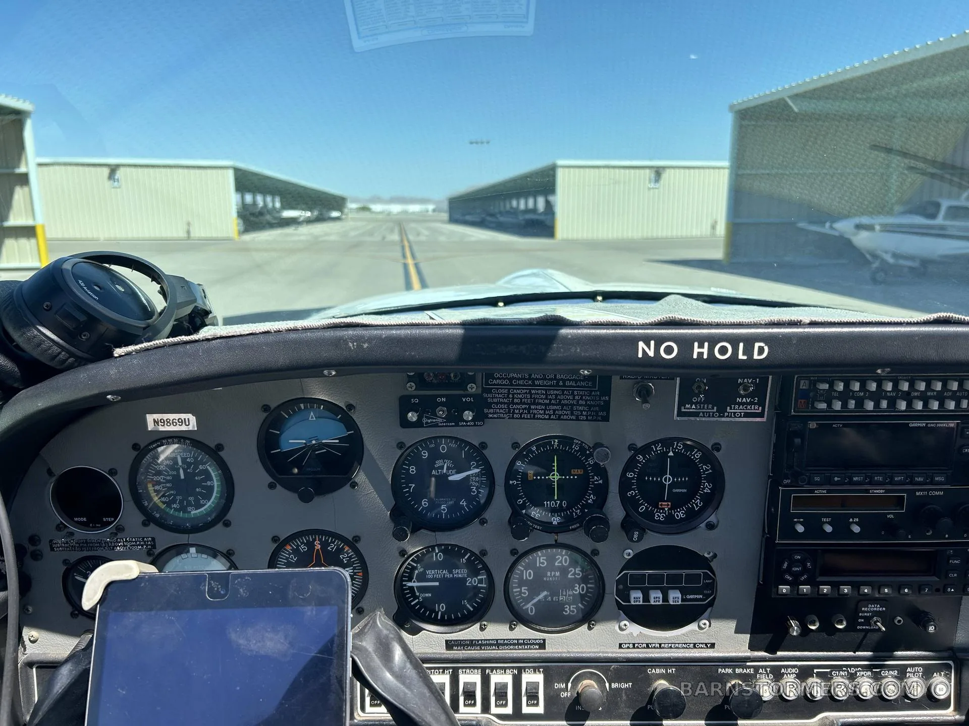 A view from the cockpit showing the instrument panel with gauges and radios, looking out towards hangars on an airfield.