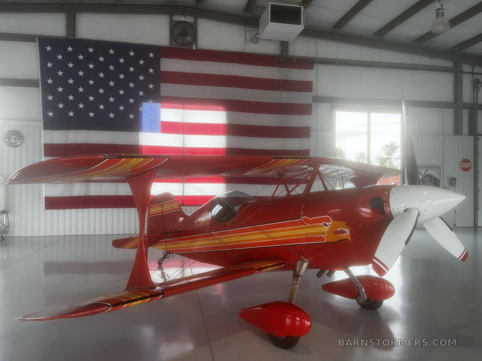 A red biplane with orange and yellow stripes and a white propeller is parked in a hangar in front of a large American flag.
