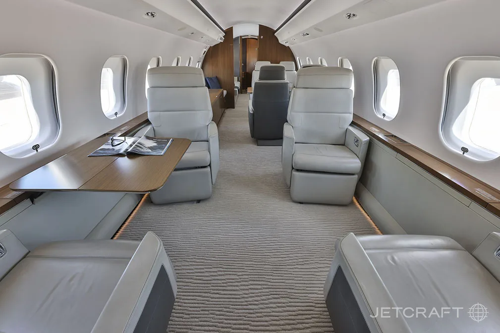 The luxurious cabin interior of a Bombardier Global 6000, featuring light grey leather seats and a wooden table.