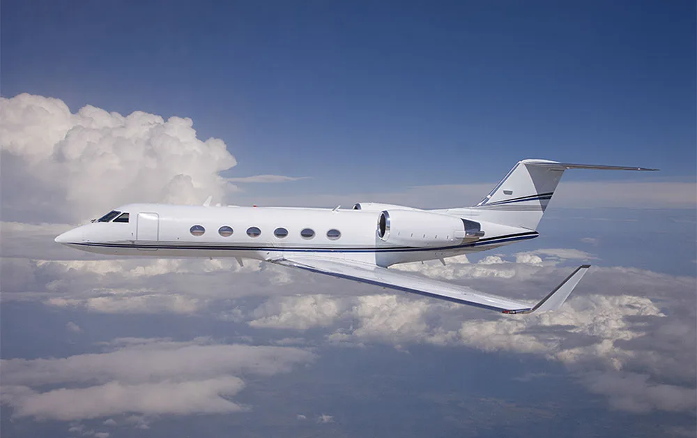A white business jet with blue accent stripes flies above a layer of clouds under a clear blue sky.
