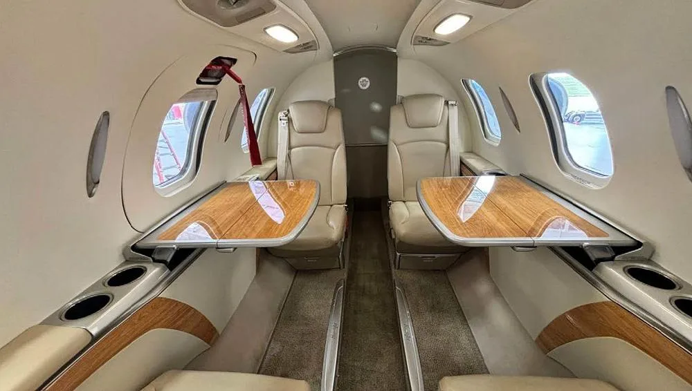 The aircraft's interior cabin features beige leather seats and two wooden fold-out tables.
